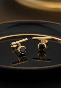 Guldtonade studs med ett horisontellt stift och en rund svart stenpendel, med en polerad yta och reflekterande detaljer.