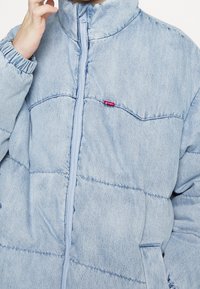 Veste rembourrée bleu clair avec une fermeture éclair, des poignets élastiques et une poche poitrine. Présente un tissu texturé, semblable au denim. Détail de petit logo.