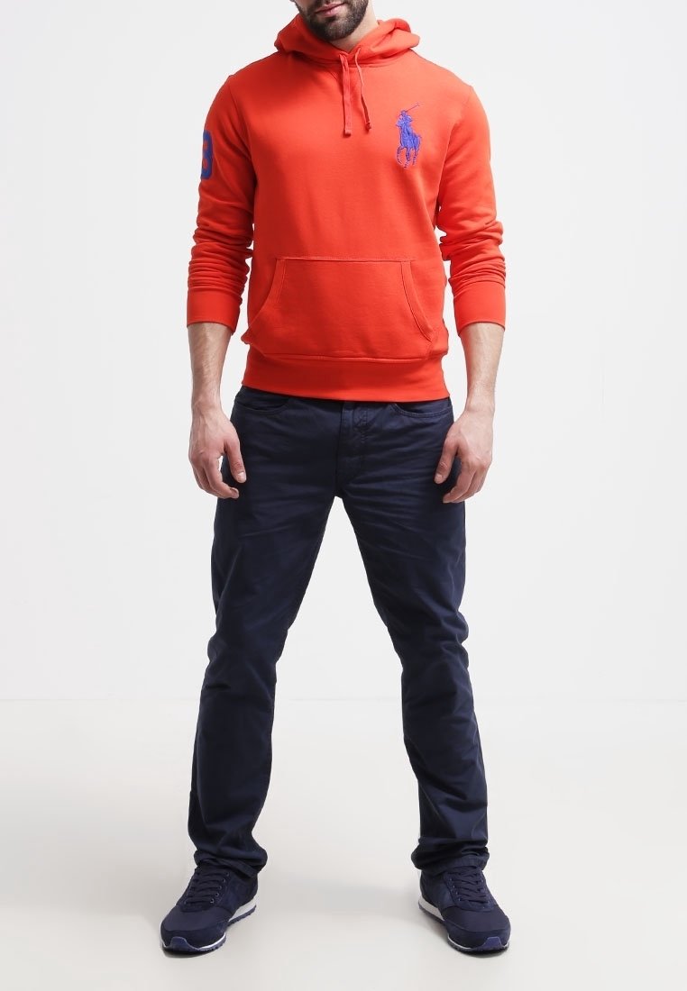 Oranger Hoodie mit einer Vordertasche, der auf der Brust ein blaues Logo trägt. Kombiniert mit marineblauen Hosen und dunklen Sneakers.