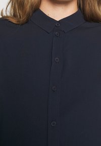Chemise boutonnée bleu marine avec un col, présentant une patte de boutonnage à l'avant avec six boutons et une texture lisse.