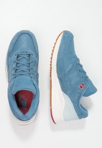 Paire de baskets en daim bleu et mesh avec semelles en caoutchouc blanches et brunes, lacets visibles, et doublure intérieure rouge, présentées vues de dessus et de côté.