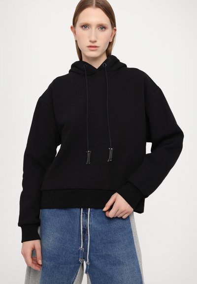 Hanorac negru, material moale, design oversized, buzunar frontal tip kangaroo, sireturi ajustabile, asortat cu blugi albaștri cu un panou lateral gri.