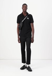 Μαύρο πόλο μπλουζάκι κοντό μανίκι σε συνδυασμό με μαύρο παντελόνι, διαθέτοντας μία μικρή τσάντα ώμου. Λείο ύφασμα, κομψή εφαρμογή και μινιμαλιστικός σχεδιασμός.