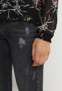 Black floral blouse with sheer fabric, featuring red and green floral patterns; paired with dark gray distressed jeans. Ruffled cuffs detail.