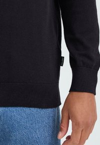 Juodas megztinis su sutrauktomis rankovėmis ir apačia. Žymė matoma kairėje pusėje. Sušalinta su mėlynomis džinsinėmis kelnėmis, parodyta ranka arti.