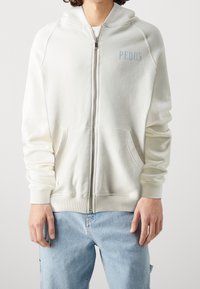 Felpa con zip di colore chiaro in tessuto morbido, caratterizzata da un cappuccio, tasche frontali e un sottile testo blu. Polsini e orlo a costine.