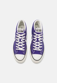 Fialové plátěné tenisky s bílými tkaničkami a gumovými podešvemi. Mají černý špičku a logo "Converse All Star" na jazyku.