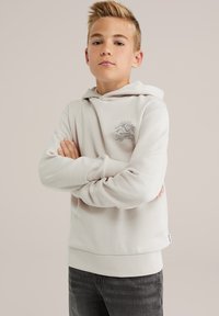 Beige hoodie met een voorzak in kangoeroestijl, ribgebreide manchetten en onderrand, met een klein grafisch ontwerp op de borst. Grijze jeans eronder gedragen.