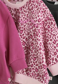 Tre barntröjor hänger, en enfärgad rosa, en rosa leopardmönstrad med "happy days," och en tredje i mörkgrå.