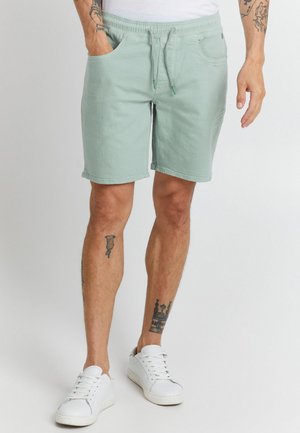 Un mannequin tatoué porte un short vert pâle à cordon et des baskets blanches, une main glissée dans une poche.