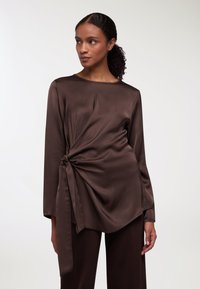 Top in satin marrone con dettaglio di nodo intrecciato in vita, maniche lunghe e scollo rotondo. Texture liscia e drappeggio fluido.