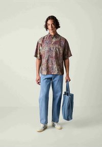 Osoba nosząca kolorową, abstrakcyjną koszulę z krótkim rękawem, jasnoniebieskie dżinsy, białe trampki, trzymająca dużą niebieską torbę w jednej ręce.