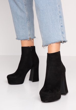 Botas de ante negras hasta el tobillo con un tacón grueso, cierre de cremallera y suela con plataforma. Combinadas con unos jeans de mezclilla cortos en un tono azul claro.