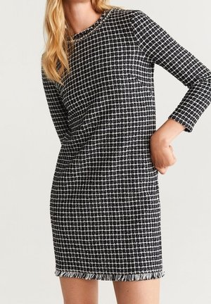 Kobieta ubrana w czarno-białą, w kratę sukienkę z długim rękawem, ozdobioną frędzlami przy dekolcie i na dole sukienki, stojąca z jedną ręką na biodrze.