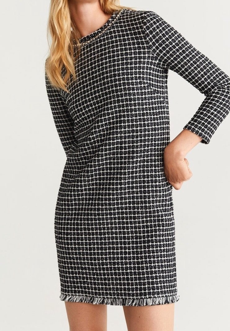 Femme portant une robe à manches longues à carreaux noirs et blancs avec des détails en franges au niveau de l'encolure et de l'ourlet, debout avec une main sur la hanche.