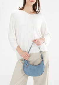 Blauwe suède handtas met een gebogen vorm, verstelbare schouderriem en ritssluiting. Model draagt een losse witte top en lichtbeige broek.