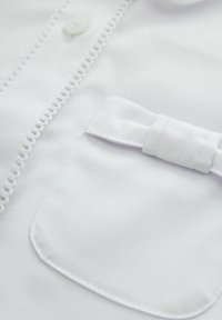 Camicia bianca in cotone con chiusura a bottoni, cuciture decorative lungo il patta e un accento a fiocco sulla tasca del petto.