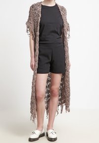 Lång, öppenstickad, brun cardigan med fransdetaljer, bärs över en svart t-shirt och shorts, i sällskap med svart och vita skor.