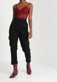 Punainen pitsinen bodysuit säädettävillä olkaimilla, korkeat mustat housut ja kiiltävät viininpunaiset nilkkurit, joissa on kärki ja paksu korko.