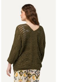 Olivgrüner Häkelpullover mit V-Ausschnitt hinten, versehen mit sechseckigen Mustern und Dreiviertelärmeln. Kombiniert mit gemusterten Hosen.