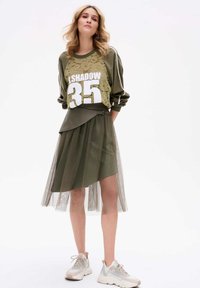 Oliwkowa bluza z koronkową nakładką i białym nadrukiem, w połączeniu z warstwową spódnicą z tiulu, dopełniona neutralnymi trampkami.