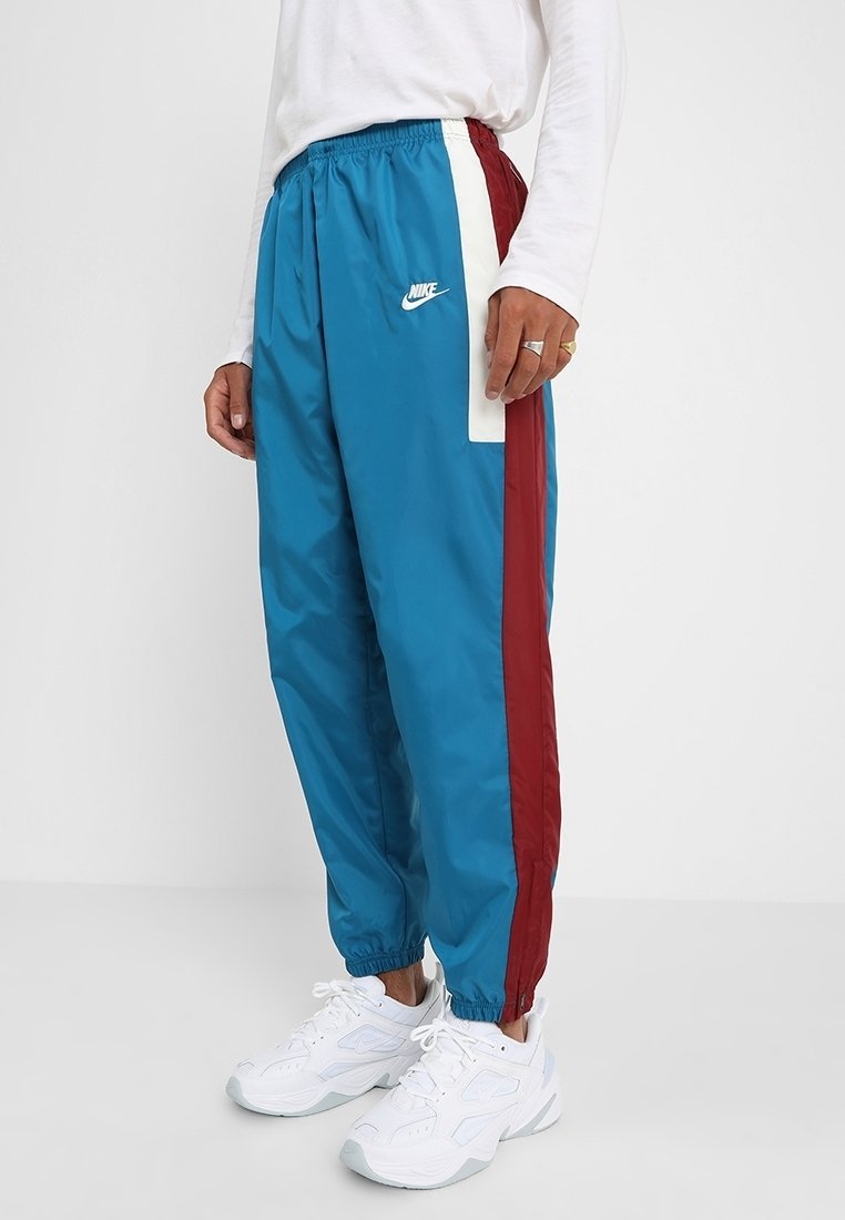 Blå Nike-träningsbyxor med vita och maroon sidosträck som bärs av en person i vita sneakers och en långärmad vit tröja.