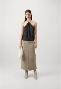 Ženska v črnem topu z ovratnikom, bež dolgi krilu, sivih gležnjarjih in z belo torbico v roki, stoji ob enobarvni svetli kulisi.