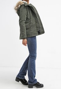 Zielono-oliwkowa parka z kapturem ozdobionym sztucznym futrem, dwoma przednimi kieszeniami i zamkiem błyskawicznym. Połączona z niebieskimi dżinsami i czarnymi butami na platformie.
