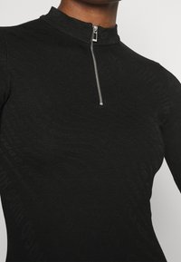 Haut noir à manches longues avec une texture discrète à motif léopard et une fermeture éclair argentée au niveau du col montant, porté par une personne à teint moyen.