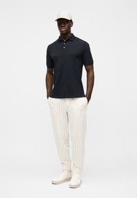 Czarna koszulka polo, kremowe spodnie w wzory, beżowa czapka i białe trampki. Casualowy fason z krótkimi rękawami i elastycznymi ściągaczami przy kostkach.