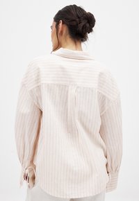Lichtroze, gestreepte blouse met kraag, lange mouwen en strikjes aan de mouwen. Beschikt over een rugplooien en een yoke-detail.