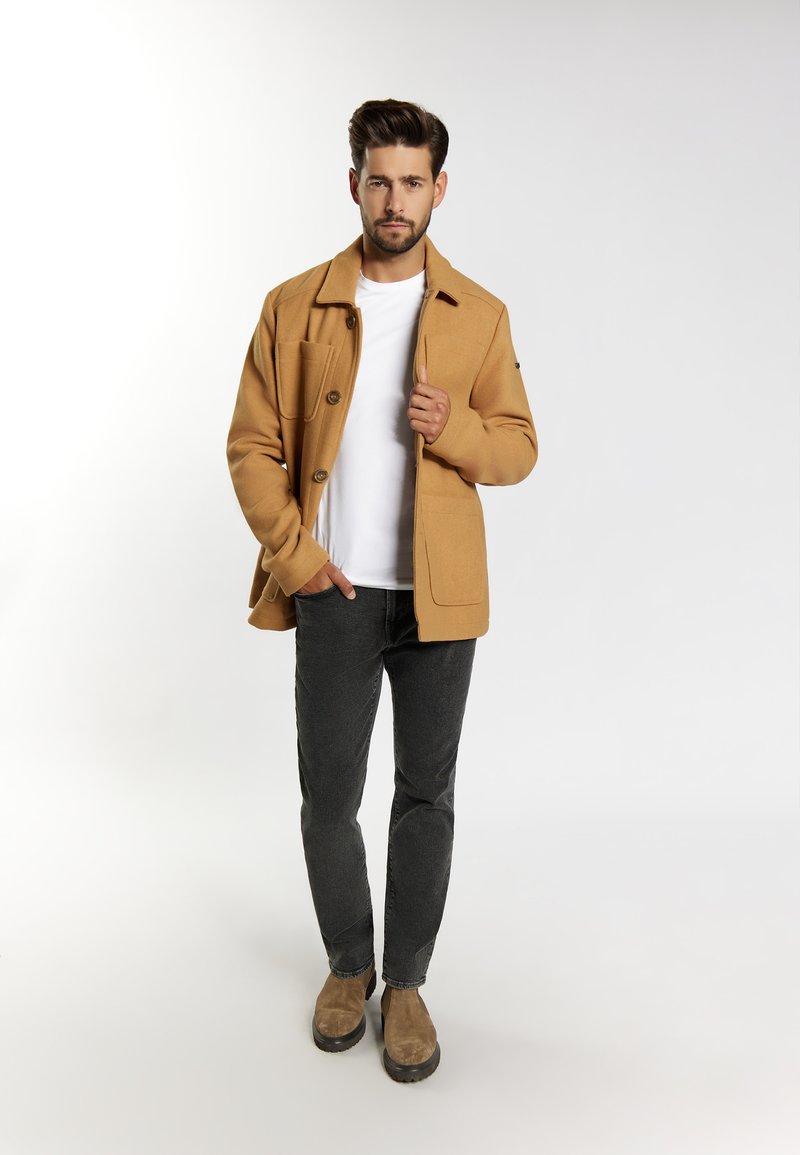 Veste en mélange de laine marron avec de grandes poches avant, fermeture à boutons, portée sur un t-shirt blanc, assortie à un jean foncé et des bottes en daim marron.