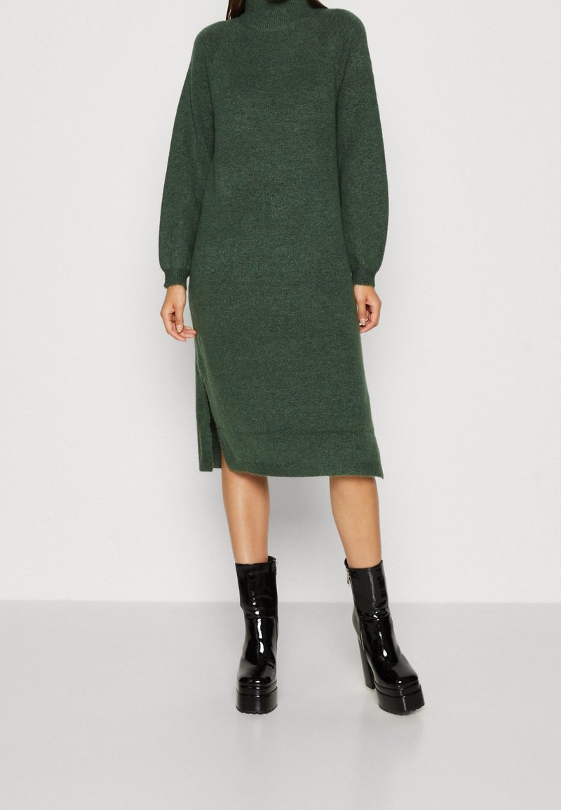 Femme portant une longue robe pull en maille vert foncé avec fente latérale et bottines à plateforme en cuir verni noir.