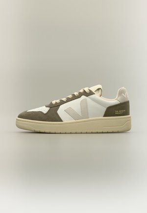 Niedriger beigefarbener und weißer Sneaker mit Wildlederdetails und "The Aegean Project" Schriftzug an der Ferse, vor neutralem Hintergrund.