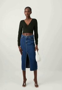 Kvinne iført en svart langermet cropped topp, blå høyt midjefed denimskjørt med frontsliss, gjennomsiktige hæler, og holder en liten hvit håndveske.