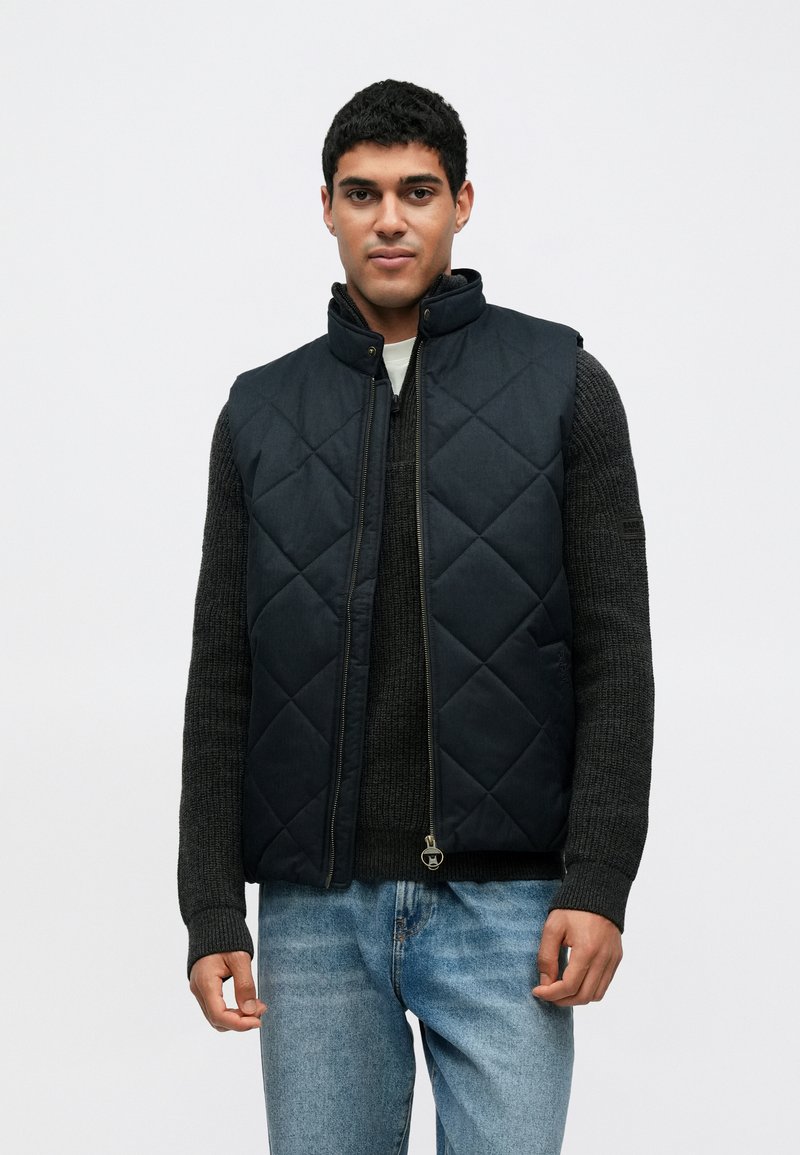 Ναυτικό μπλε γιλέκο καπιτονέ με ψηλό γιακά, κλείσιμο με φερμουάρ και διακριτική λεπτομέρεια λογότυπου. Φορεμένο πάνω από σκούρο γκρι πουλόβερ, συνδυασμένο με ανοιχτό μπλε τζιν.