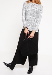 Schwarze Handtasche mit doppelten Griffen, strukturiertem Stoff und kastenförmigem Design. Tragen mit einem grauen gemusterten Pullover und weiten schwarzen Hosen, kombiniert mit schwarzen Stiefeln.