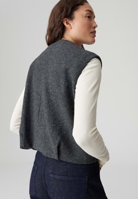 Gilet in lana grigia con design corto, senza maniche e finitura testurizzata. Indossato sopra una maglia a maniche lunghe a coste color crema e denim scuro.