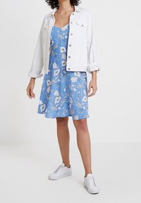 Vestido floral azul con patrones blancos, con corsé ajustado y falda acampanada; combinado con una chaqueta de mezclilla blanca y zapatillas blancas.