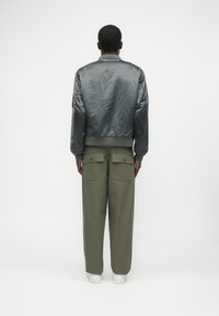 Casaco bomber cinza em cetim com punhos canelados, combinado com calças largas verde-oliva com dois bolsos traseiros e sapatilhas brancas.