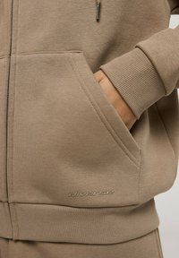 Beżowa bluza z kapturem na zamek błyskawiczny, z ściągaczami przy rękawach i u dołu. Posiada kieszeń z przodu oraz wyszywane logo "diverse" z boku. Gładka tekstura materiału.