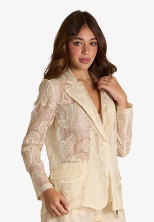 Donna con lunghi capelli castani che indossa un blazer in pizzo crema trasparente con motivi paisley, tenendo il colletto con una mano.