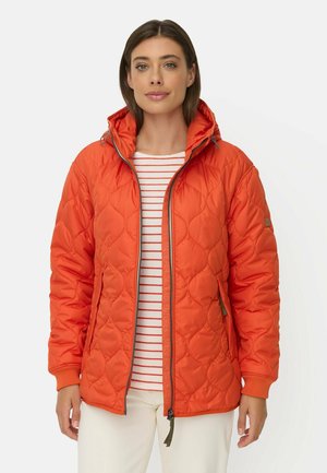 Kvinde iført en orange quiltet jakke over en hvid og orange stribet skjorte med hvide bukser, stående foran en ensfarvet hvid baggrund.