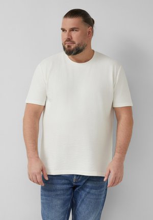 Man with beard and tied-back hair wearing a textured white short-sleeve shirt and blue jeans against a plain gray background.