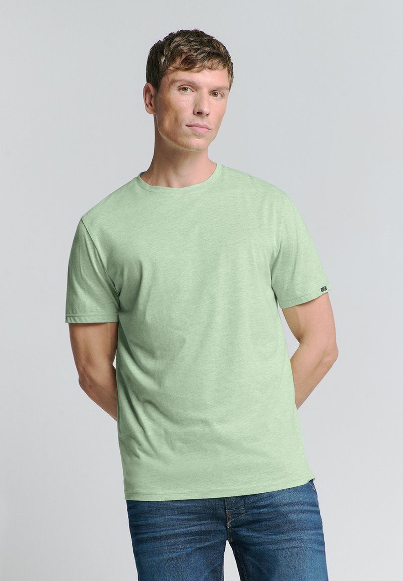 Lichtgroene t-shirt met korte mouwen van zachte stof, met een ronde hals en een klein logo op de linker mouw. Draagt met blauwe jeans.