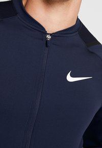 Marineblå atletisk top med fuld lynlås, med en buet krave og teksturerede detaljer. Fremtrædende hvid Nike swoosh-logo på brystet.