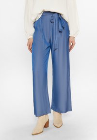 Wijdvallende broek in een gladde, lichtblauwe stof met een hoge tailleband en bijpassende strikceintuur; gedragen met beige haklaarzen.
