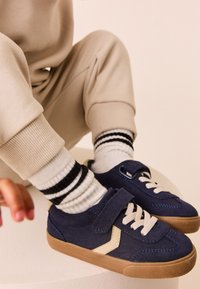 Baskets bleu marine avec une bride en velcro, une semelle contrastante couleur tan, et des lacets blancs. Portées avec des chaussettes rayées blanches et noires.