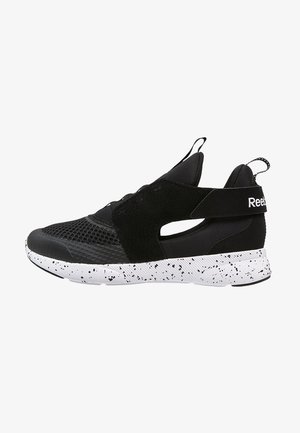 Zapatilla deportiva negra con parte superior de malla y tejido, suela blanca gruesa con manchas negras, correa elástica en el mediopié y detalle del logo.