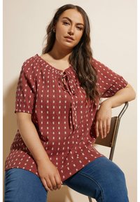 Blusa rossa con texture, maniche corte, dettaglio a chiave con lacci e motivi geometrici bianchi, abbinata a jeans in denim blu.
